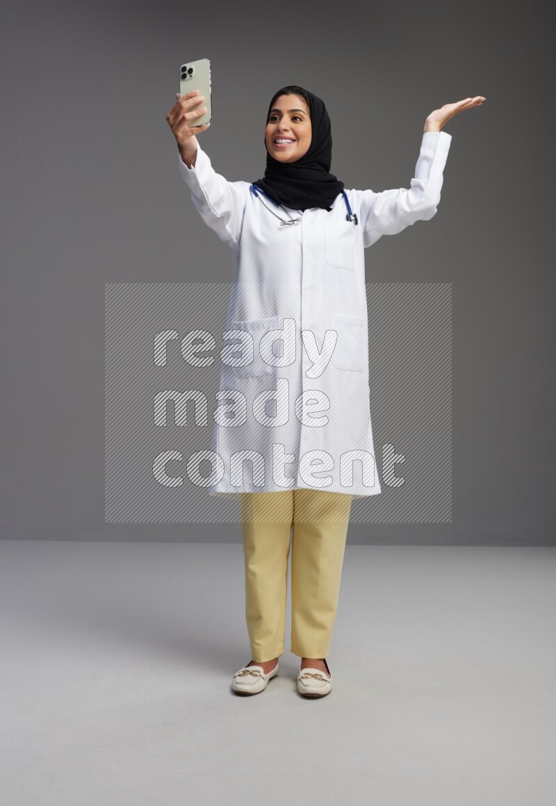 Saudi woman wearing lab coat with stethoscope standing taking selfie on Gray background