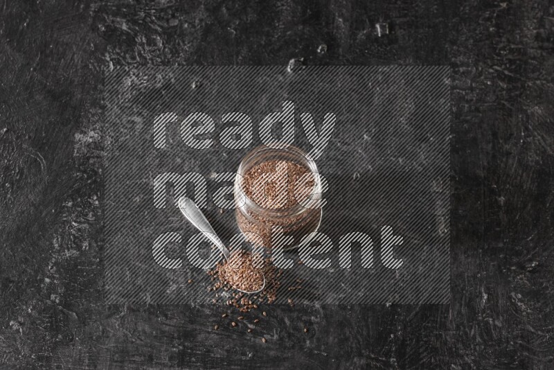 A glass jar full of flaxseeds with a metal spoon full of the seeds on a textured black flooring