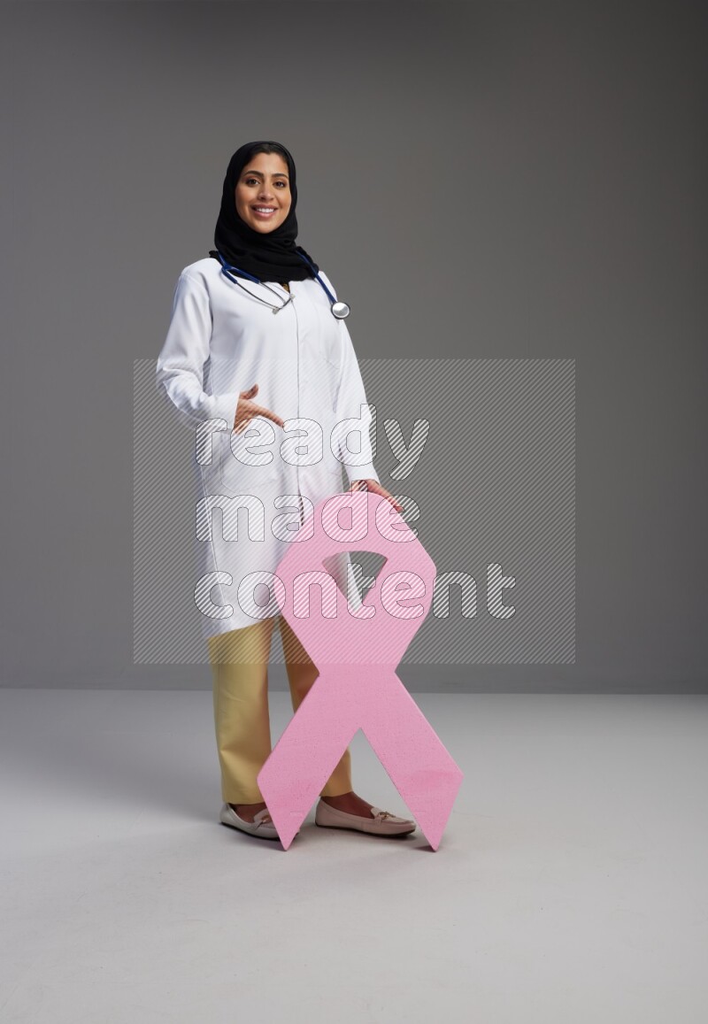 Saudi woman wearing lab coat with stethoscope standing holding awareness ribbon symbol standing on Gray background
