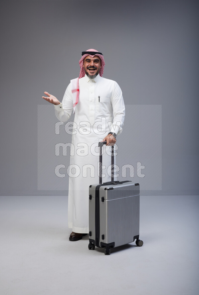 Saudi man wearing Thob and red Shomag standing holding Travel bag on Gray background