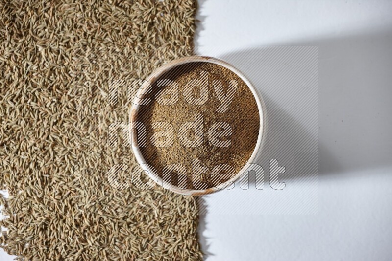 وعاء بيج ممتلئ ببودرة الكمون مع حبوب الكمون تحت الوعاء على خلفية بيضاء