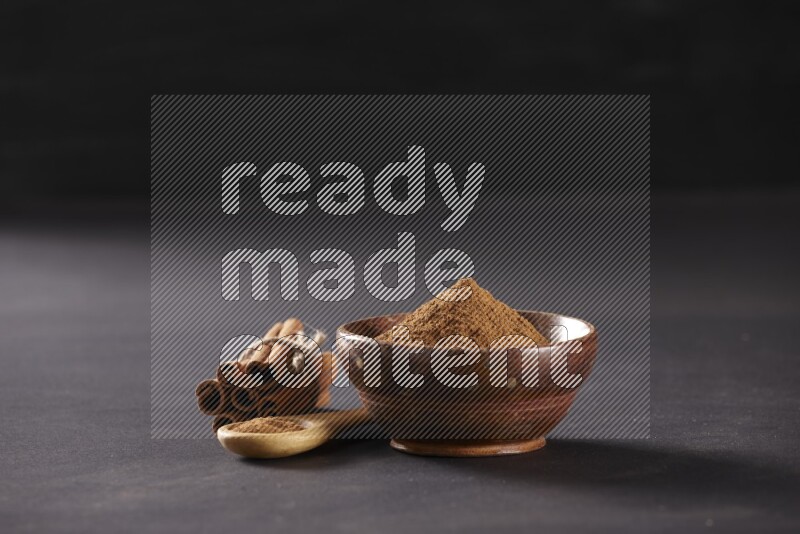 Cinnamon sticks stacked and bounded beside a wooden bowl full of cinnamon powder and a wooden spoon full of powder on black background