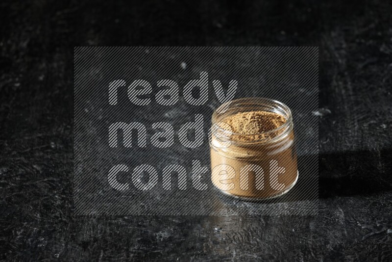 A glass jar full of allspice powder on a textured black flooring