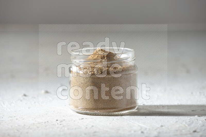 A glass jar full of cumin powder on textured white flooring