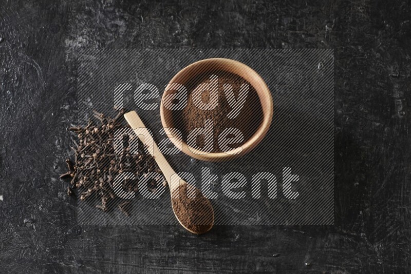 A wooden bowl and a wooden spoon full of cloves powder with spreaded cloves on a textured black flooring