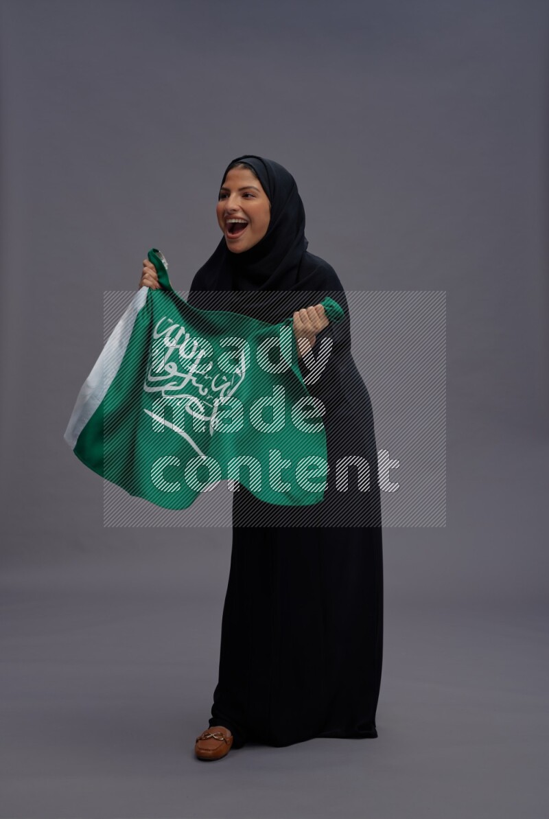 Saudi woman wearing Abaya standing holding Saudi flag on gray background