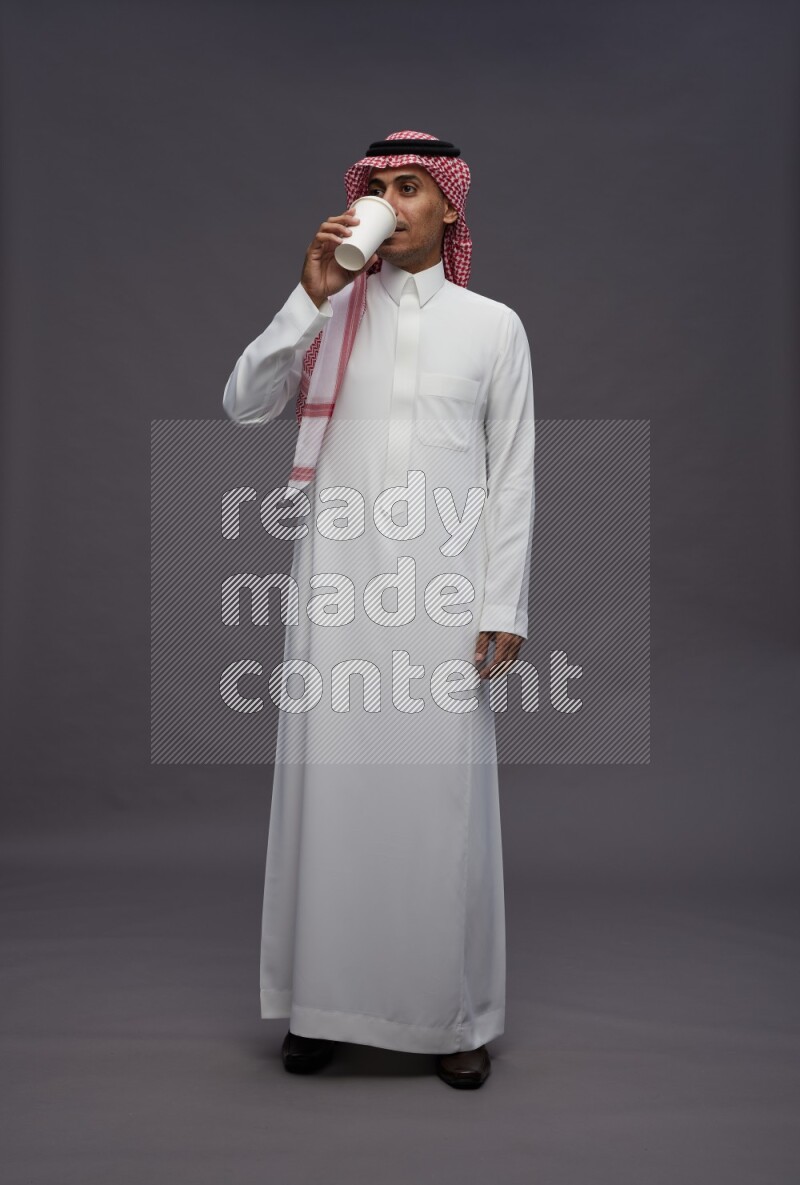 Saudi man wearing thob and shomag standing holding paper cup on gray background