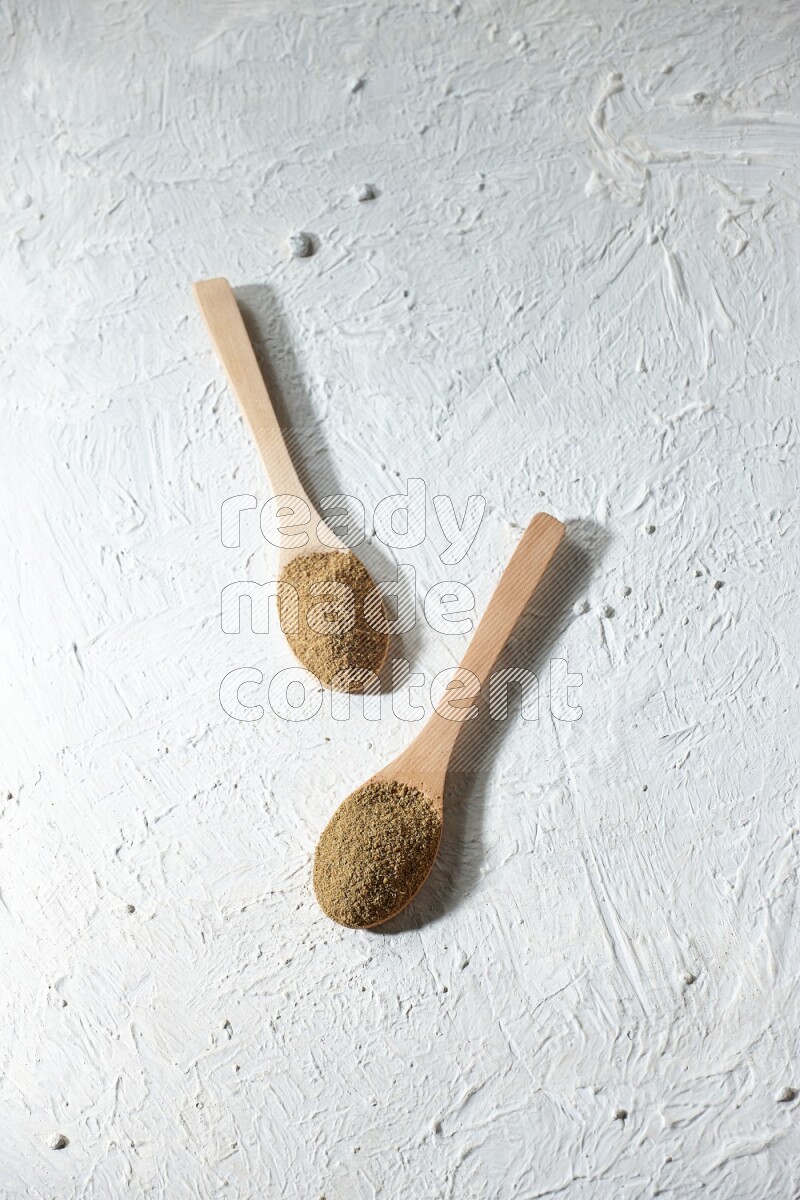 2 Wooden spoons full of cumin powder on a textured white flooring