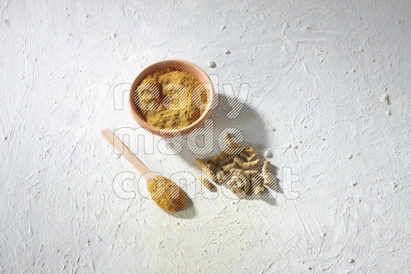 A wooden bowl and wooden spoon full of turmeric powder with dried turmeric fingers on textured white flooring