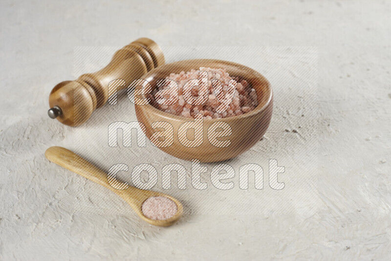 A wooden bowl and spoon filled with coarse pink himalayan salt and a wooden grinder beside them on white background