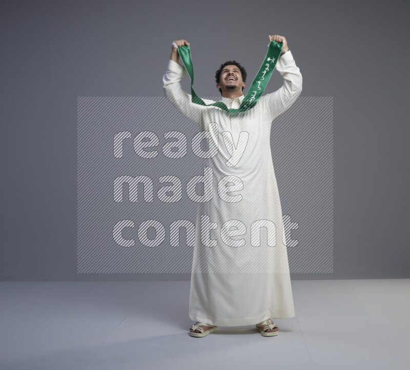 A Saudi man standing wearing thob holding Saudi flag scarf on gray background