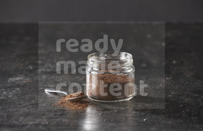 A glass jar full of cloves powder with a metal spoon on a textured black flooring