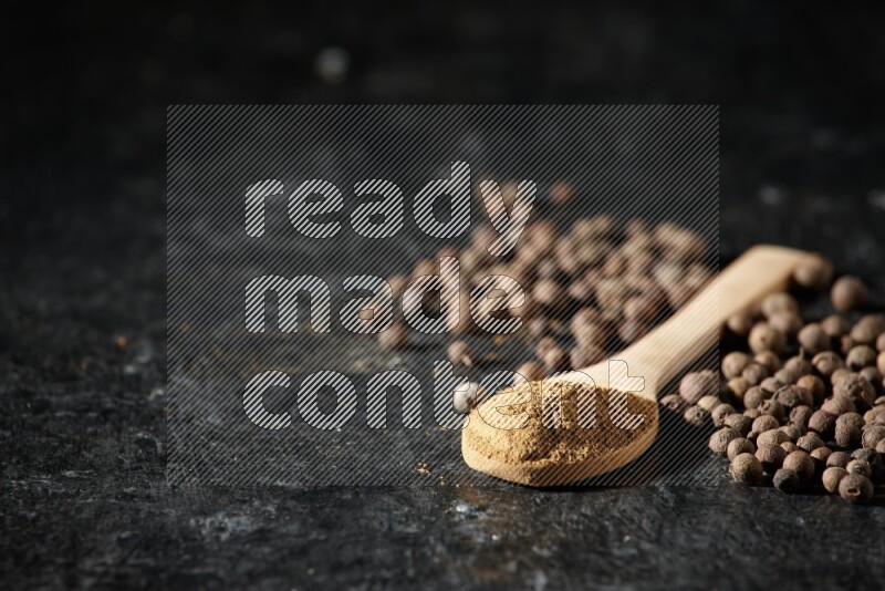Wooden spoons full of all spice powder and allspice whole balls beside it on a textured black flooring