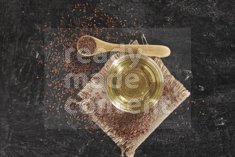 A glass bowl full of flaxseeds oil and wooden spoon full of flaxseeds with seeds spread on burlap fabric on a textured black flooring