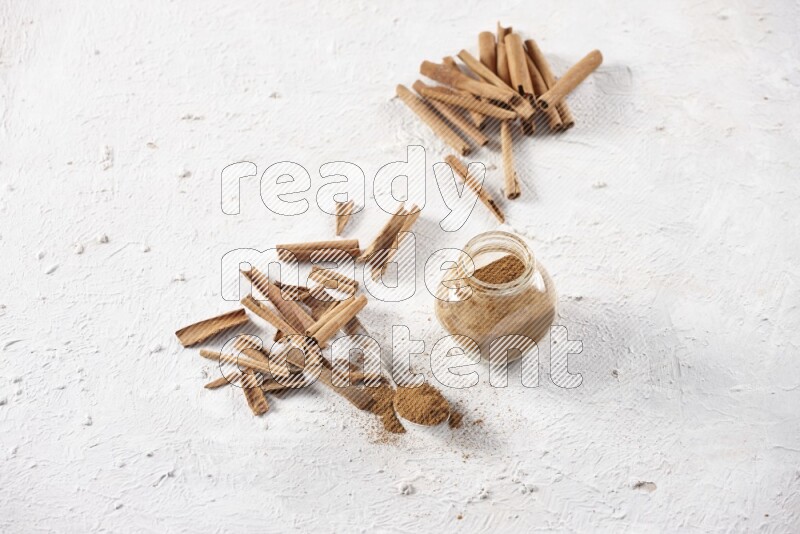 Herbal glass jar full cinnamon powder and a metal spoon surrounded by cinnamon sticks on a white background