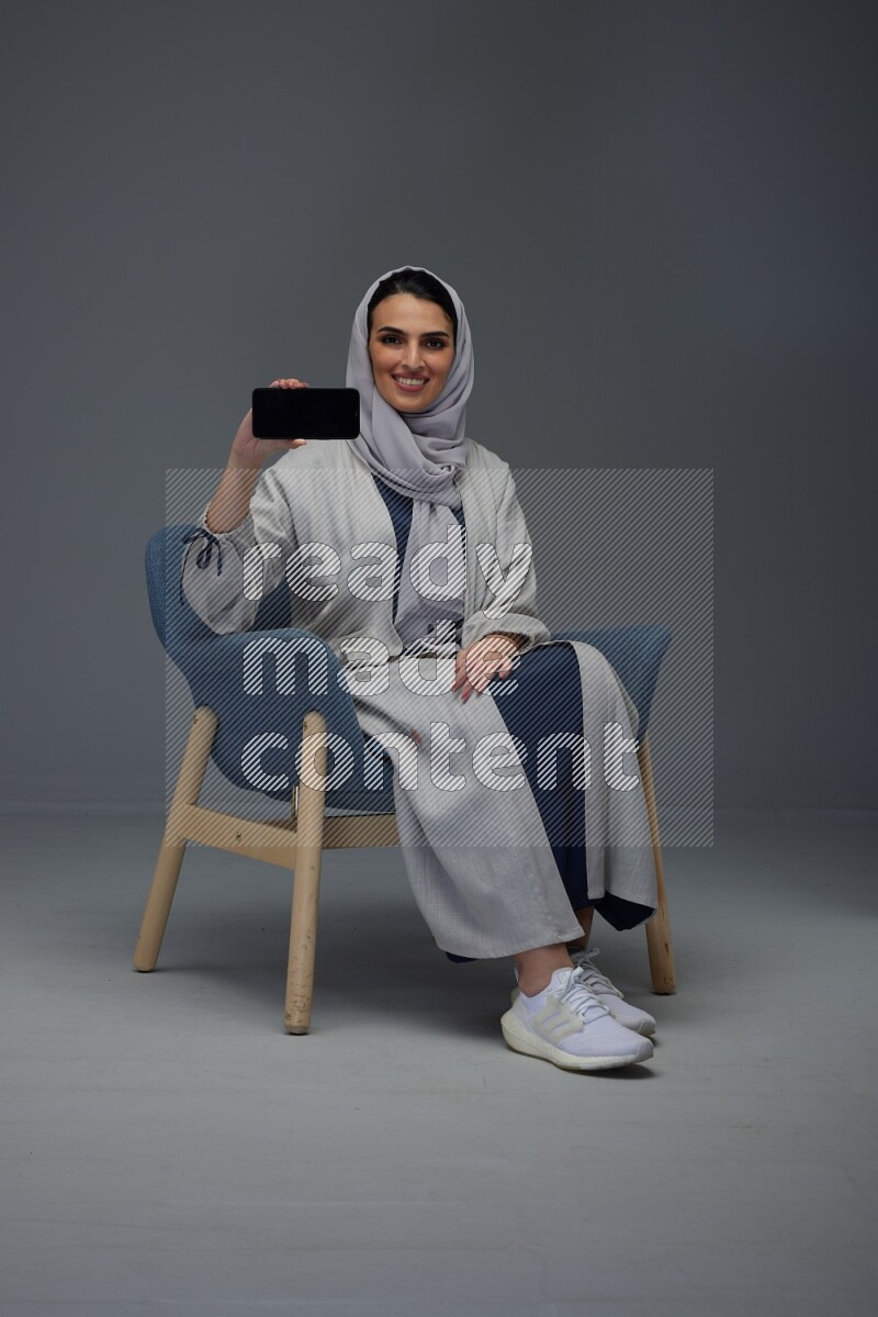 A Saudi woman wearing a light gray Abaya and head scarf sitting on a dark grey chair and showing the phone's screen eye level on a grey background