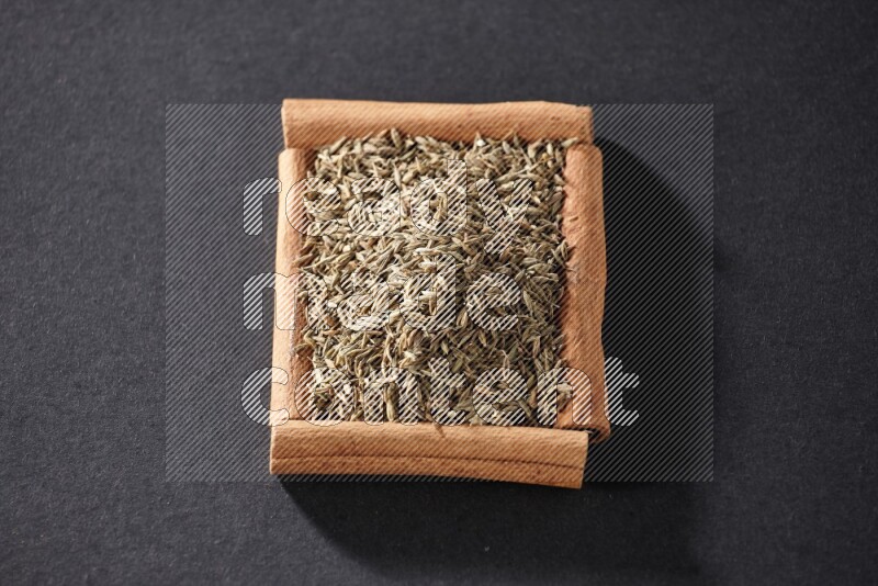 أربعة من أعواد القرفة مكونة شكل مربع بداخلها حبوب الكمون علي خلفية سوداء