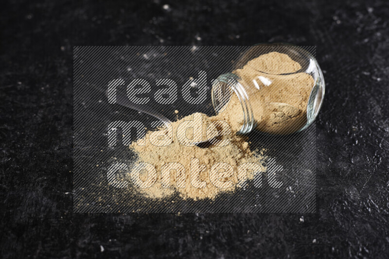 A glass jar full of ground ginger powder flipped with some spilling powder on black background