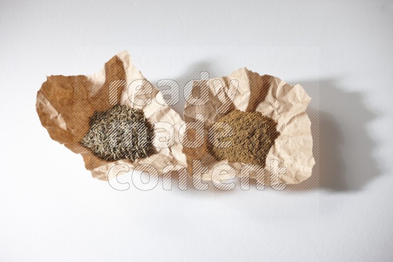 Cumin seeds and cumin powder in 2 crumpled pieces of paper on white flooring