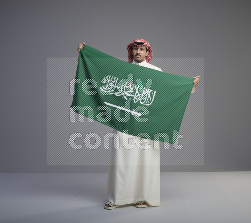 A saudi man standing wearing thob and red shomag holding big saudi flag on gray background