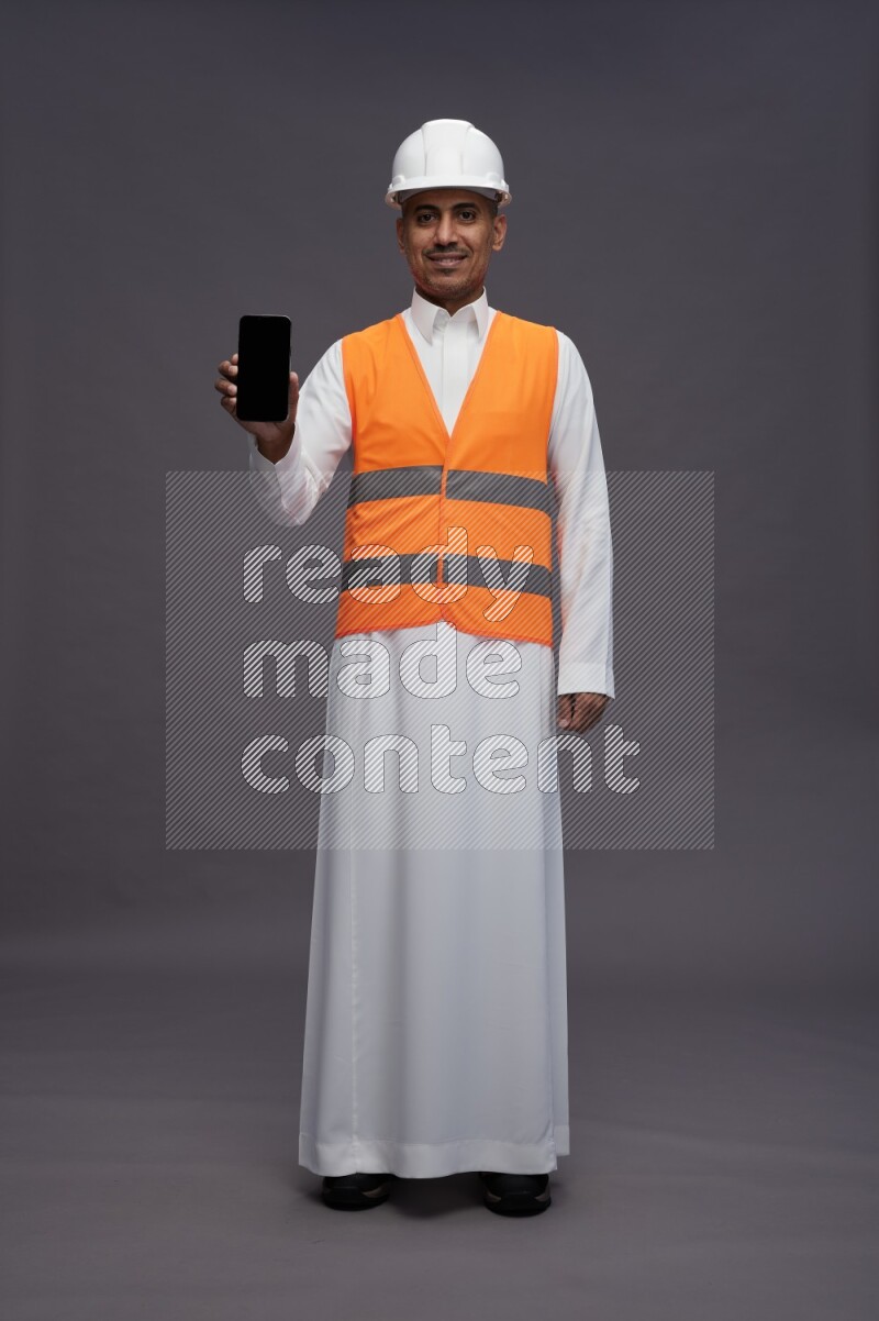Saudi man wearing thob with engineer vest standing showing phone to camera on gray background