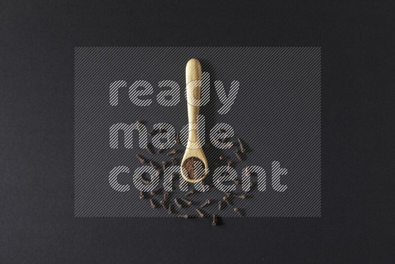 ملعقة خشبية ممتلئة ببودرة القرنفل مع حبوب القرنفل المتناثرة على خلفية سوداء