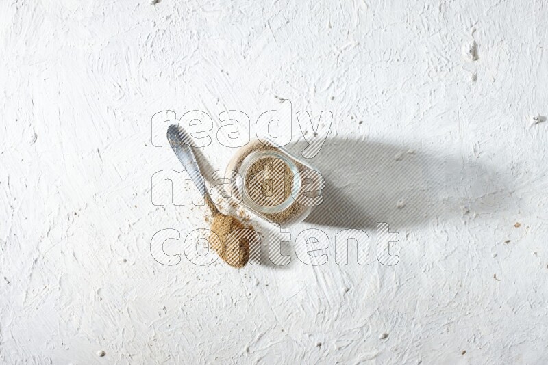 A glass spice jar and metal spoon full of cumin powder on textured white flooring