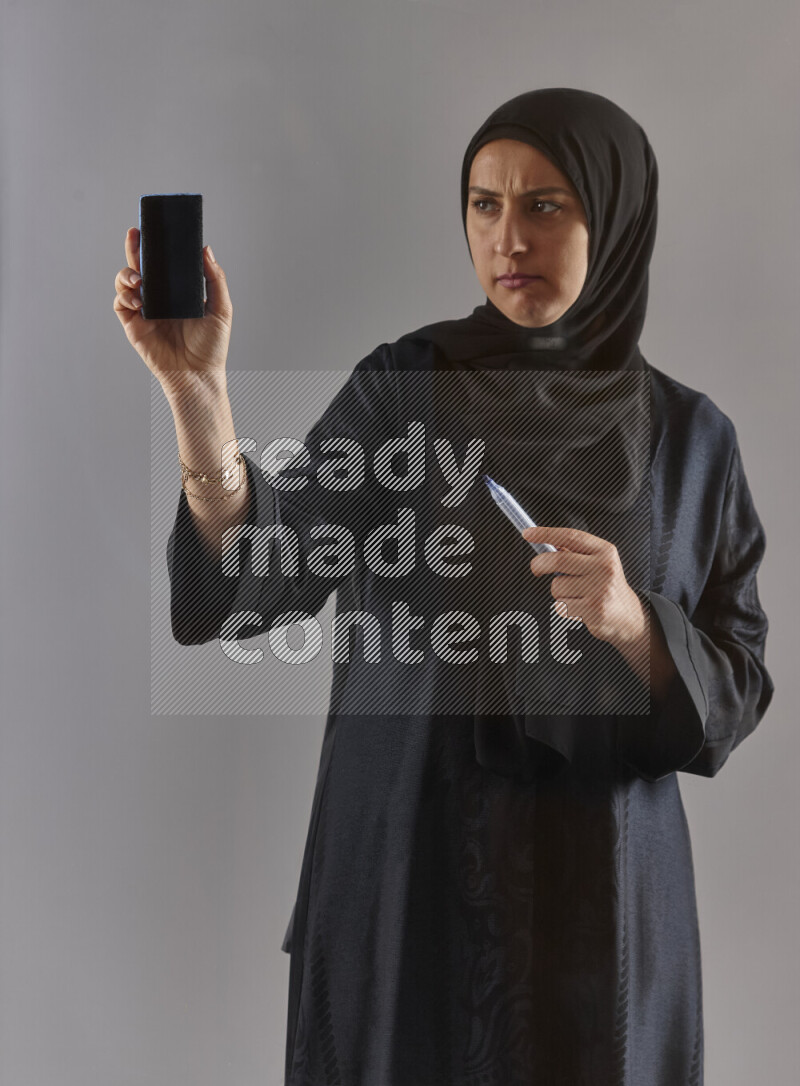 Woman in black abaya and hijab holding a marker pen to write on a transparent board with different reactions and expressions on grey background