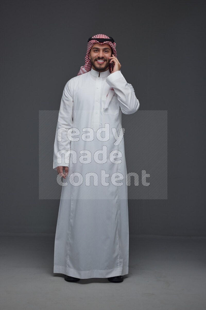A Saudi man wearing Thobe and Shmagh talking in the phone eye level on a gray background