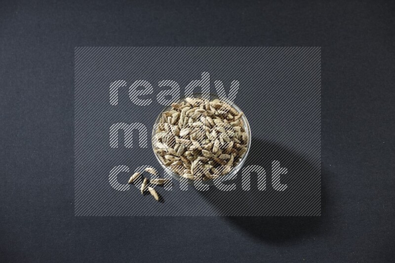A glass bowl full of cardamom and more seeds spreaded beneath the bowl on black flooring