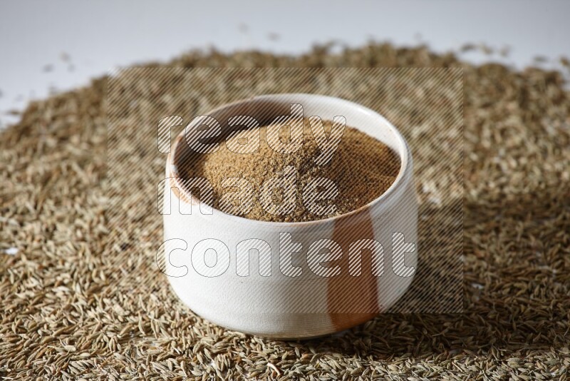 A beige bowl full of cumin powder with cumin seeds beneath it the bowl on a white flooring