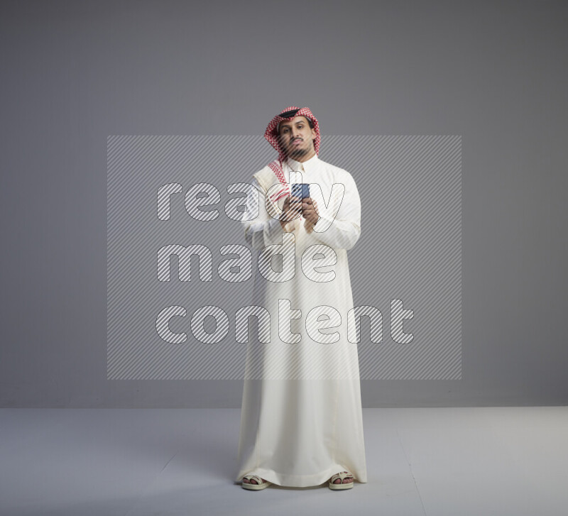 A  Saudi man standing wearing thob and red shomag texting on phone on gray background
