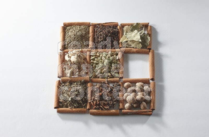 9 squares of cinnamon sticks full of cardamom in the middle surrounded by nutmeg, cinnamon, bay laurel leaves, cloves, cumin, dried ginger, dried basil and star anise on white flooring