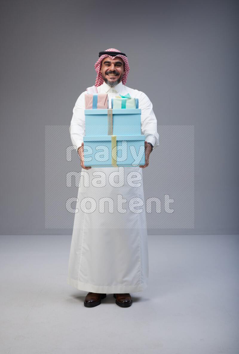 Saudi man Wearing Thob and red Shomag standing holding gift box on Gray background