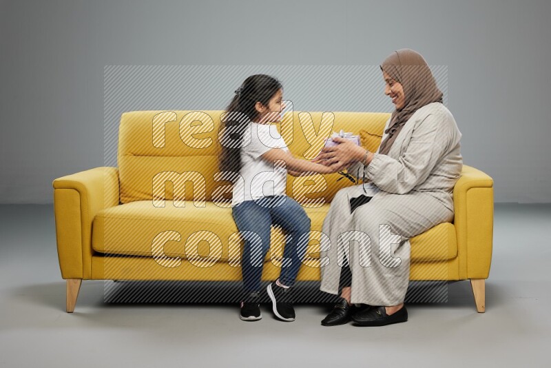 A girl sitting on a yellow sofa and giving a gift to her mother on gray background