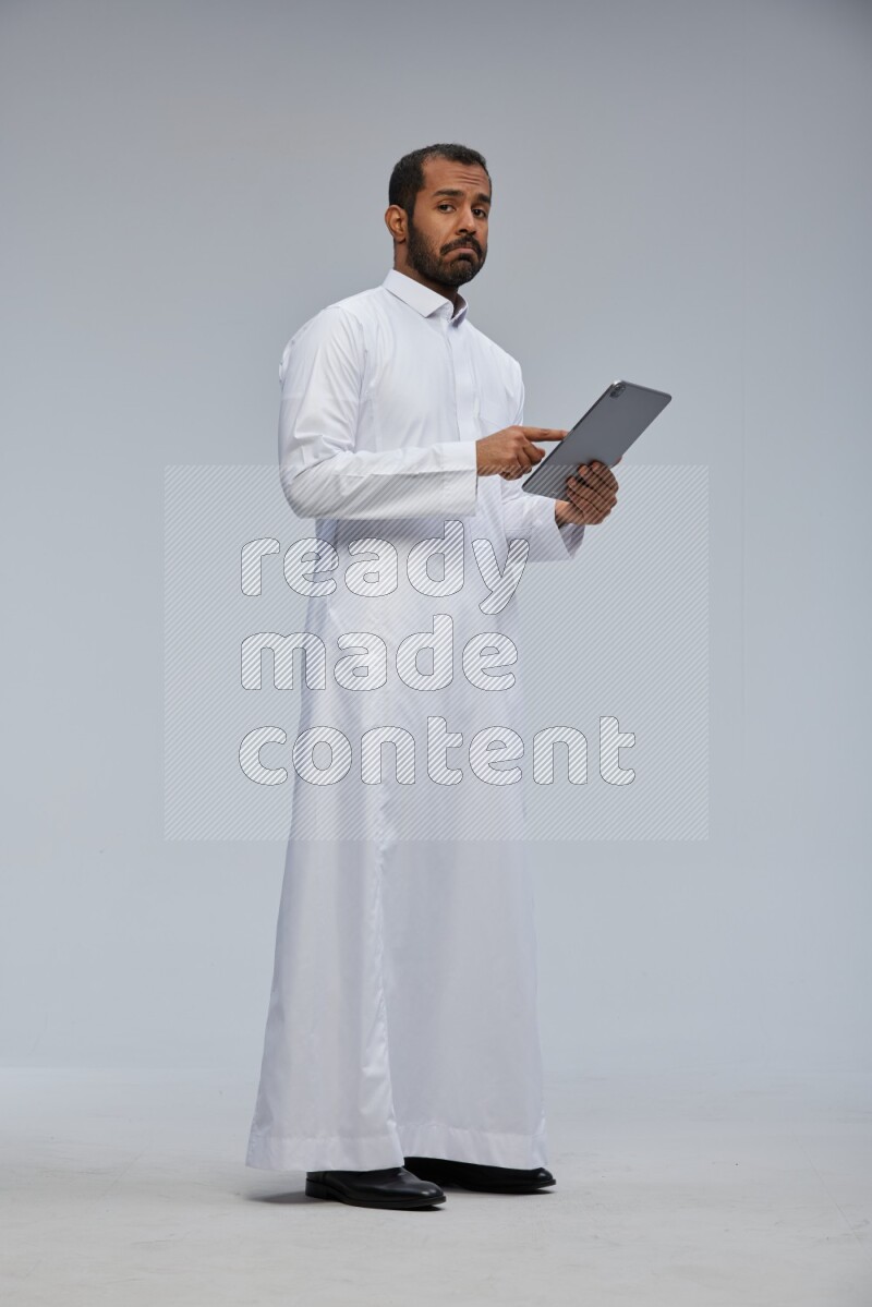 Saudi man wearing Thob standing working on tablet on Gray background
