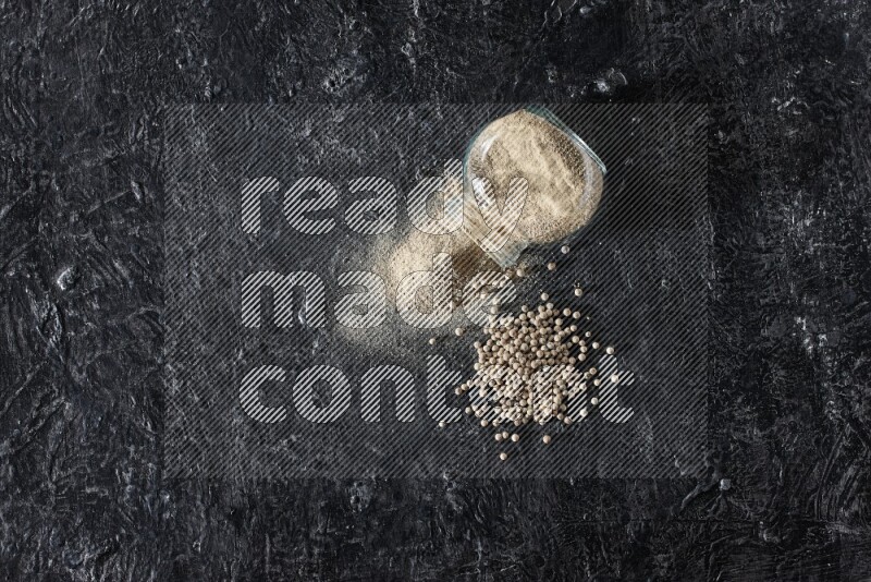 A flipped herbal glass jar full of white pepper powder with spilled powder and pepper beads on textured black flooring
