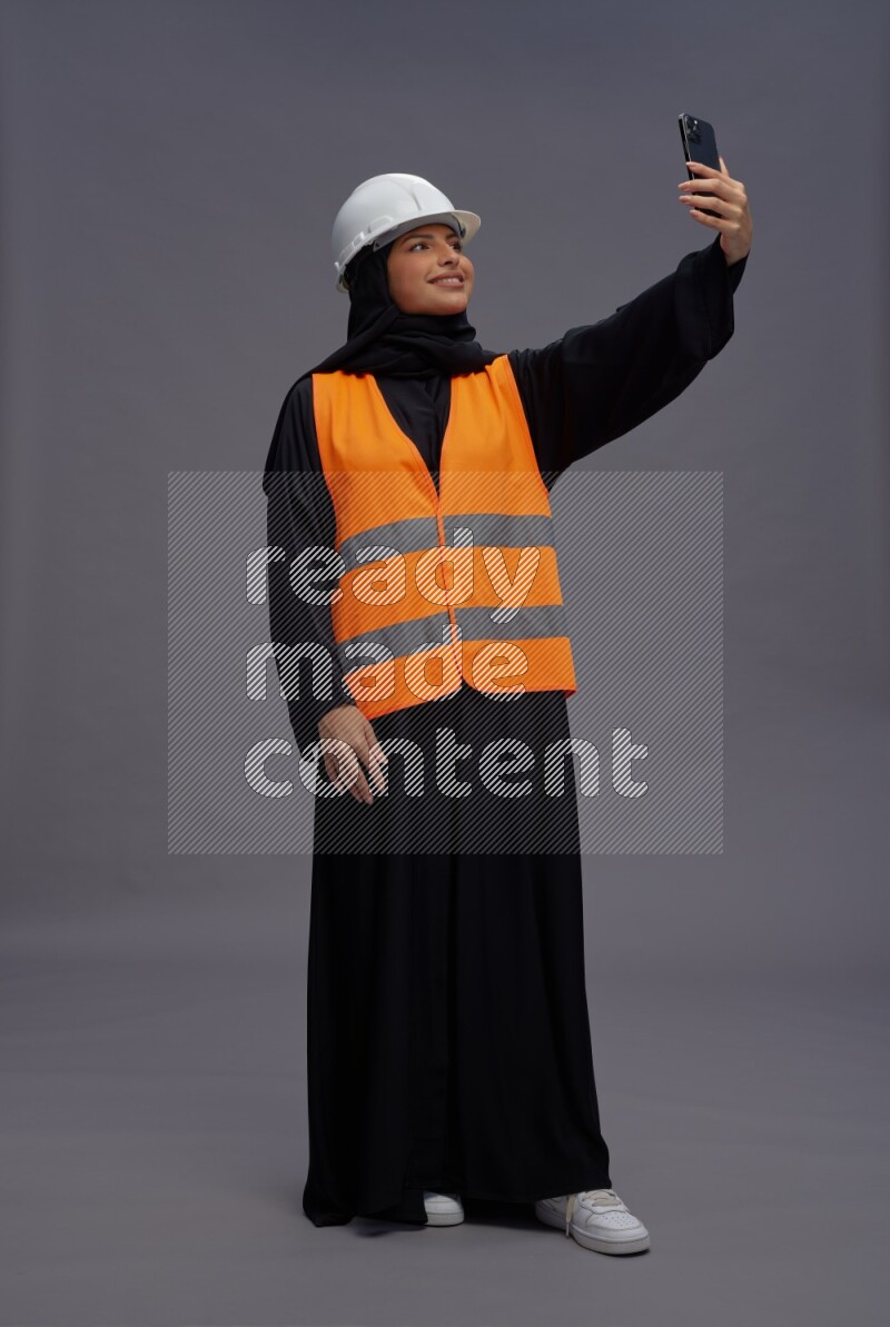 Saudi woman wearing Abaya with engineer vest standing taking selfie on gray background