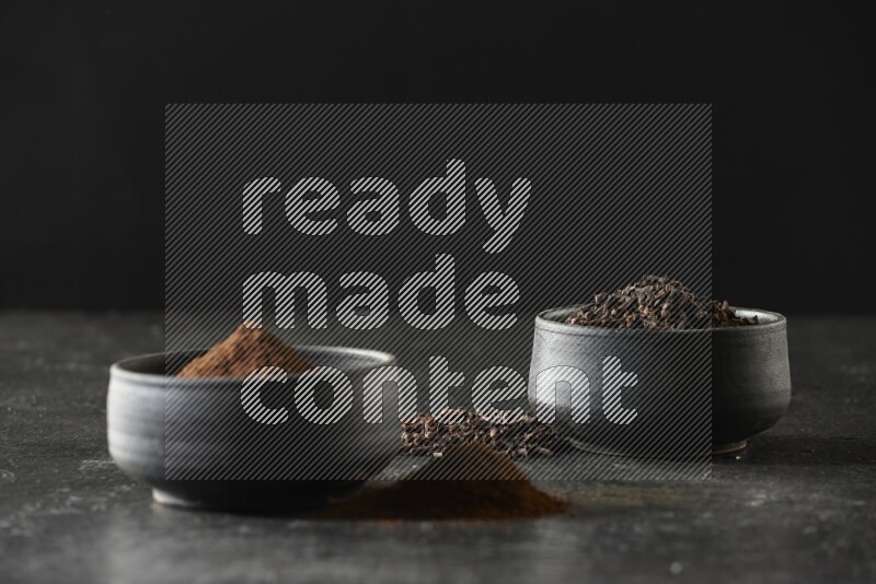 2 Black pottery bowls full of cloves and the other full of cloves powder on textured black flooring
