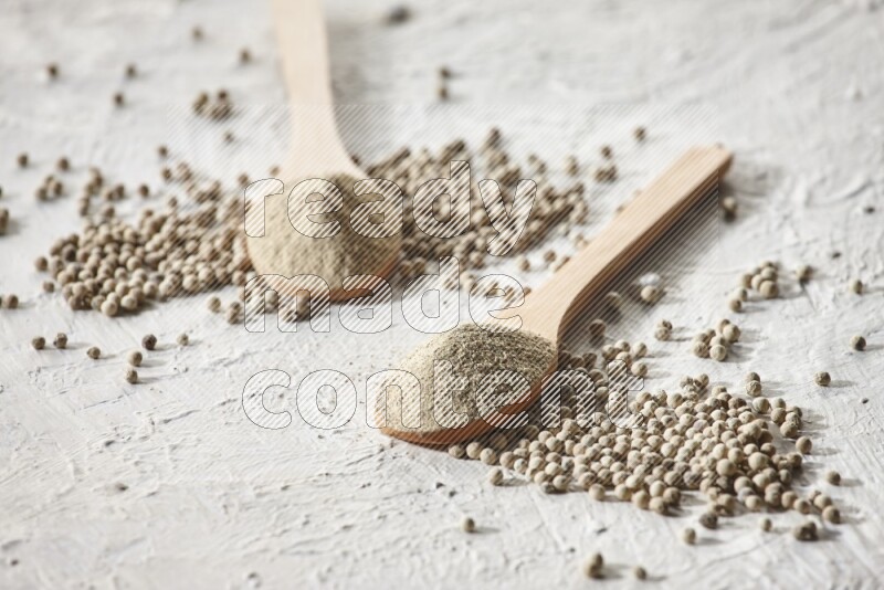 2 wooden spoons full of white pepper powder with white pepper beads on textured white flooring