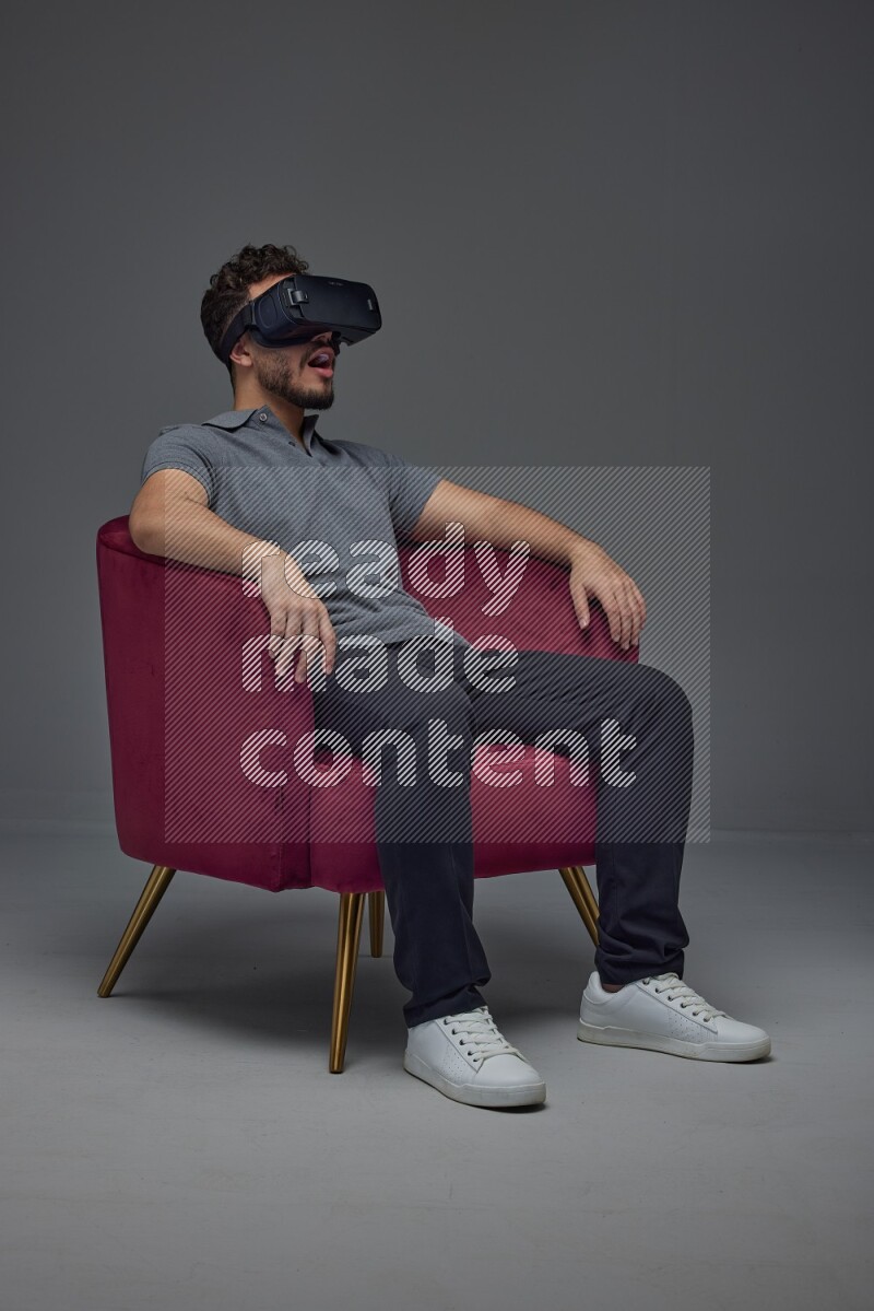 A man wearing casual and wearing VR while sitting on a burgundy chair eye level on a gray background