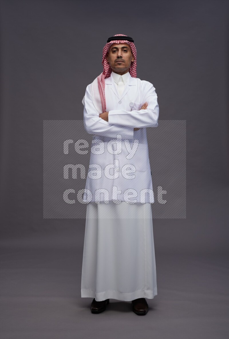 Saudi man wearing thob with lab coat and shomag with pocket employee badge standing with crossed arms on gray background