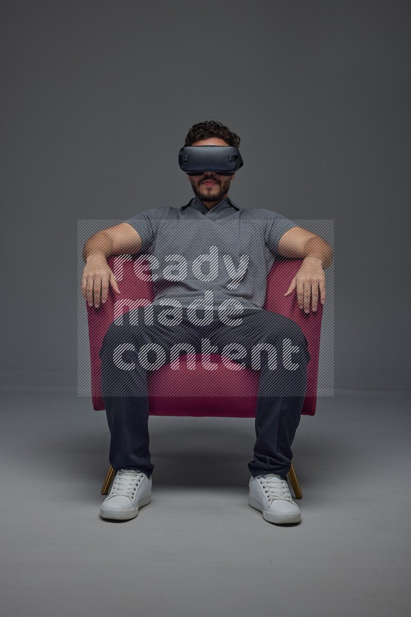 A man wearing casual and wearing VR while sitting on a burgundy chair eye level on a gray background