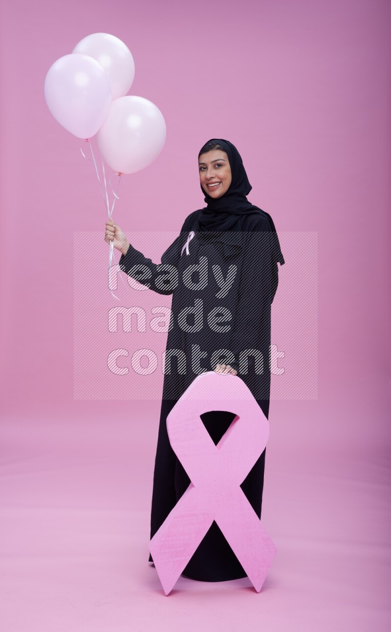 Saudi woman wearing pink ribbon on Abaya standing holding balloons and awareness sign on pink background