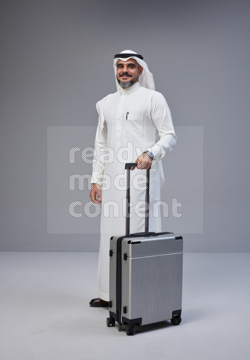 Saudi man wearing Thob and white Shomag standing holding Travel bag on Gray background