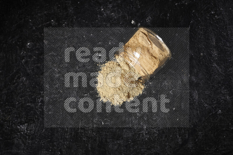 A glass jar full of ground ginger powder flipped with some spilling powder on black background