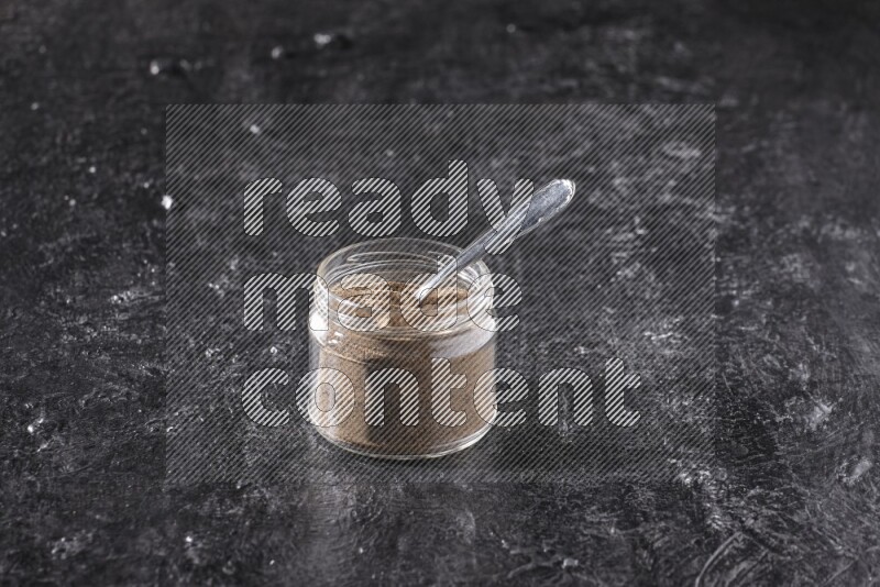 A glass jar full of black pepper powder and a metal spoon on a textured black flooring