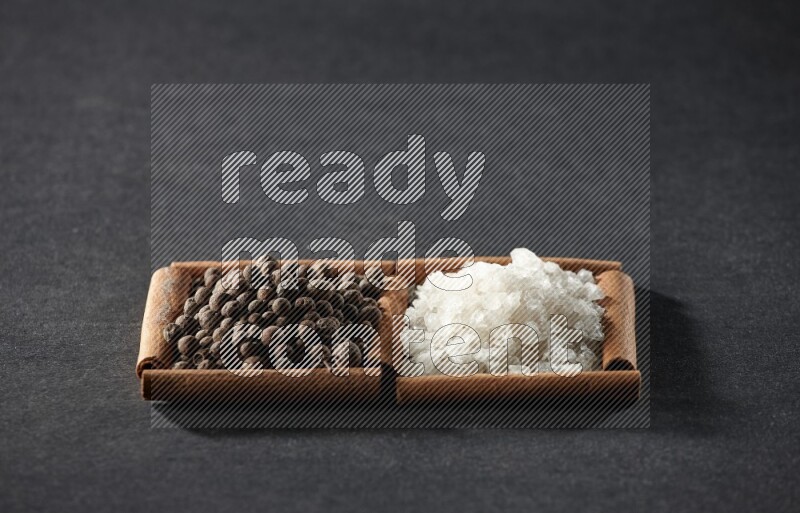 مربعان من أعواد القرفة ممتلئان بالملح الأبيض وحبوب الفلفل الحلو علي خلفية سوداء