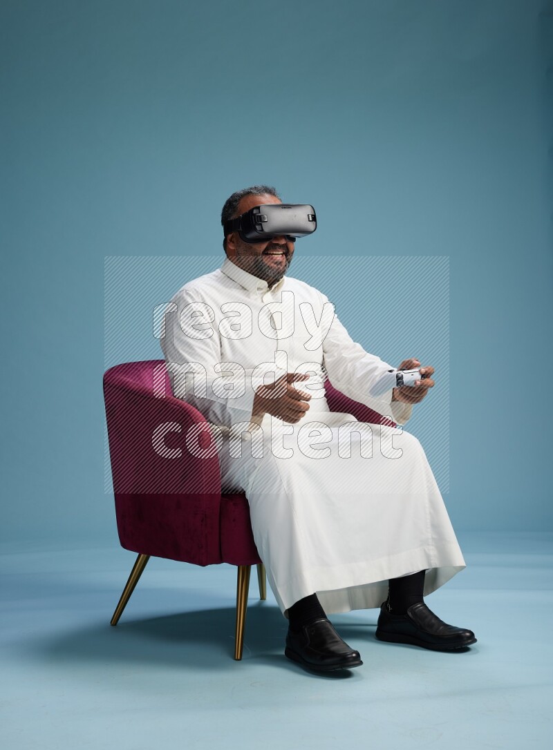 Saudi Man without shimag sitting on chair  gaming with VR and joystick on blue background