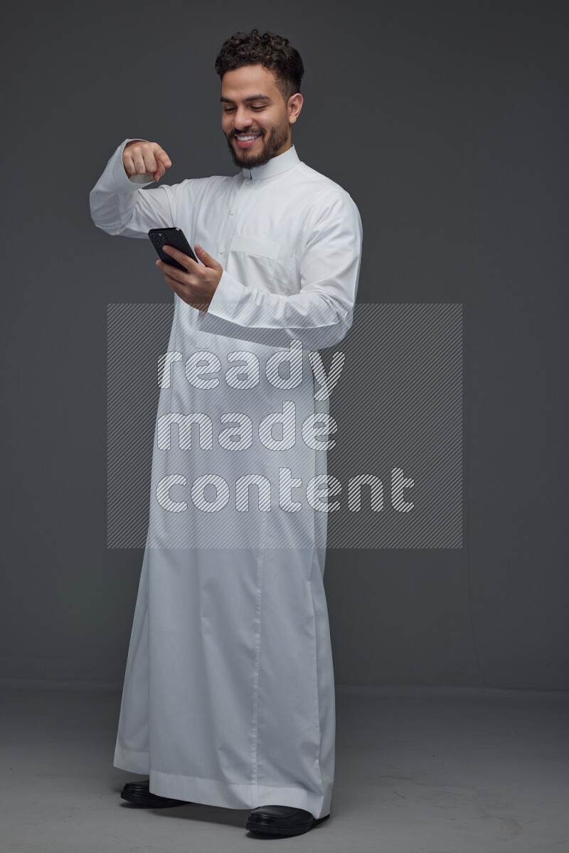 A Saudi man wearing Thobe and holding his phone while pointing to it eye level on a gray background